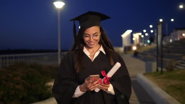 College examen flicka promenader med diplom leende och använda sin smartphone. — Stockvideo