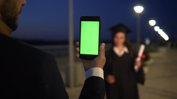 Homem segurando o telefone com tela verde como se estivesse tirando uma foto de uma estudante graduada que está de pé na frente dele. Foto para memórias. — Vídeo de Stock