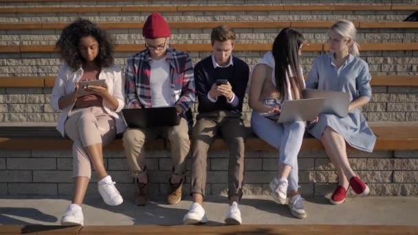 Multi-etnische studenten zitten in een rij opgenomen door telefoons, computers, tabs. Apparaatverslaving. — Stockvideo