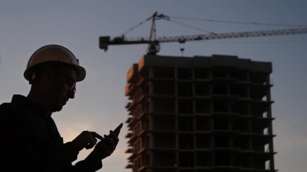 Ingeniero en el sitio de construcción usando su teléfono enviando mensajes de texto a algo. Silueta. — Vídeos de Stock