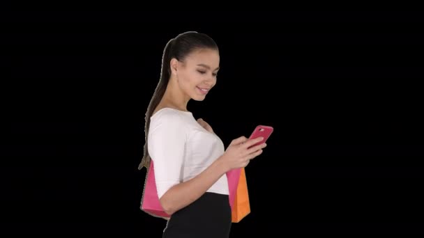 Hermosa joven usando un teléfono móvil caminando y sosteniendo bolsas de compras, Alpha Channel — Vídeo de stock