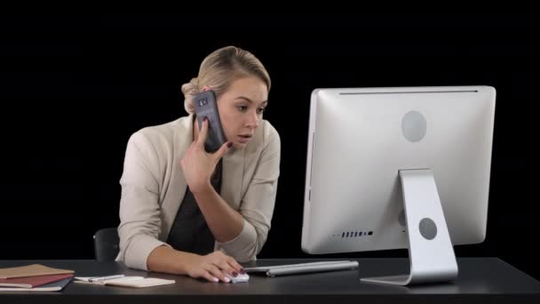 Приваблива молода жінка говорити по мобільному телефону і посміхаються при сидячи на її робоче місце в офісі і, дивлячись на комп'ютер, Альфа-канал — стокове відео
