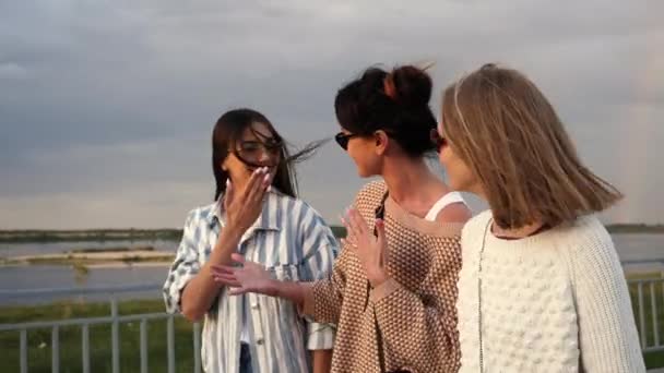 Tres mujeres jóvenes en gafas de sol caminan por el paseo marítimo y chismorrean . — Vídeos de Stock