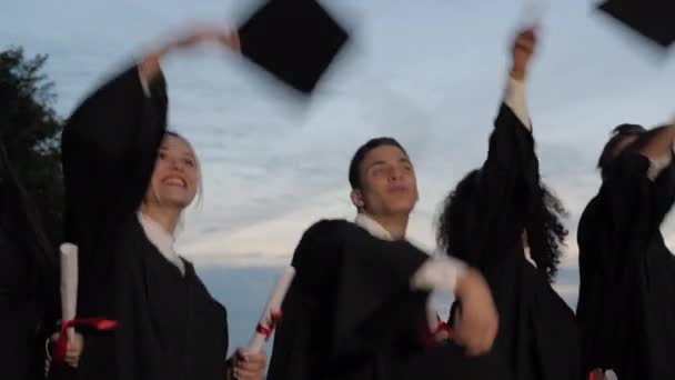 Felice ed eccitato gruppo etnico misto di amici studenti il giorno della laurea lanciare cappelli in aria mentre si cammina .. — Video Stock