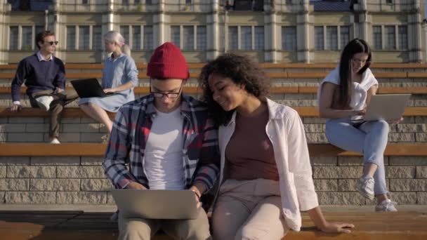Casual koppel surfen op het internet met behulp van laptop buiten. Studenten doen projecten op de campus. — Stockvideo