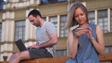 Diz üstü bilgisayar kullanan bir adam ve akıllı telefon kullanan bir kadın açık hava bankında oturuyor..