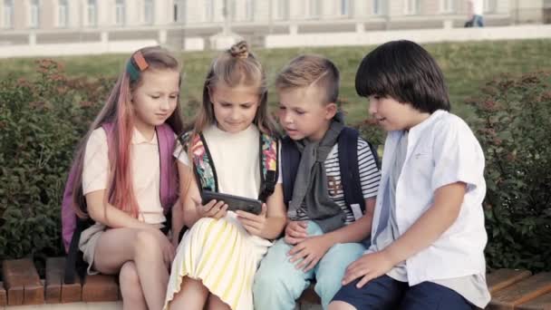 公園に座ってスマートフォンを見ている4人のガジェット中毒の子供. — ストック動画
