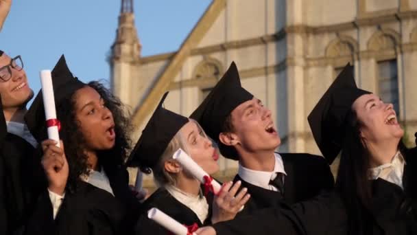 Corridas mistas amigos graduados em vestidos tradicionais pretos e bonés fazendo selfies no smartphone . — Vídeo de Stock