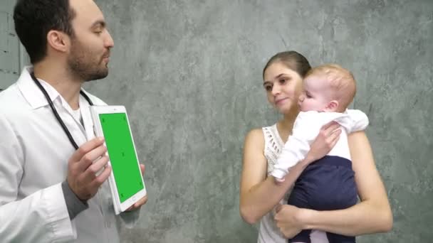 医師は母親と赤ん坊の少年にタブレットで医療結果を示しています。緑の画面のモックアップ表示. — ストック動画