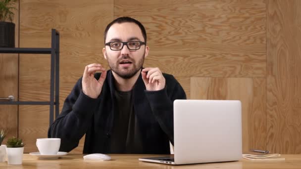 Joven hombre hipster barbudo sentado en su escritorio con el ordenador portátil hablando con la cámara . — Vídeos de Stock