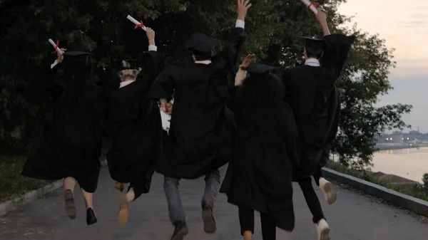 Vijf jonge afgestudeerden springen omhoog tijdens het lopen. — Stockfoto