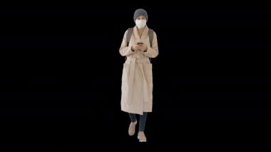 Paltolu bir kadın tıbbi maske takıyor telefon kullanıyor ve yürüyor, Alpha Channel 'da.