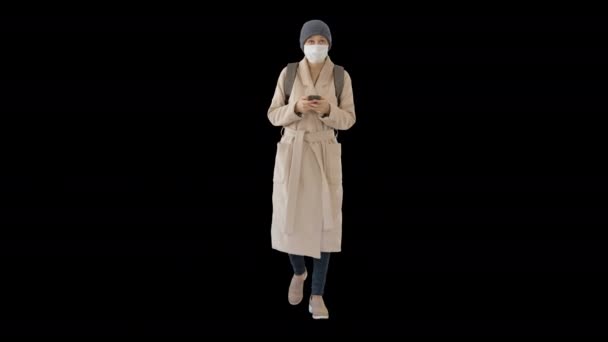 Woman dressed in a coat wearing medical mask using phone and walking, Alpha Channel — Stock Video