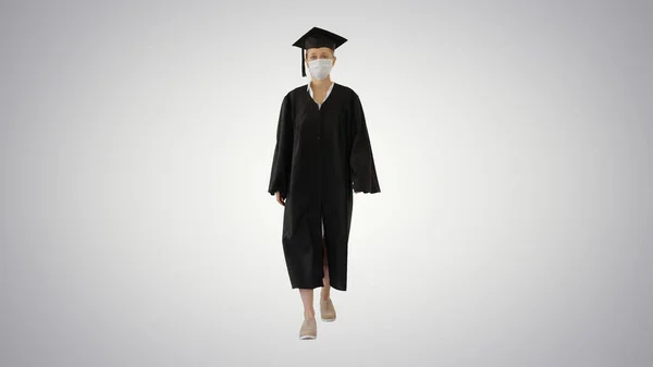 Vrouwelijke afgestudeerde in Cap and Gown dragen medische masker Wandelen op gradiënt achtergrond. — Stockfoto