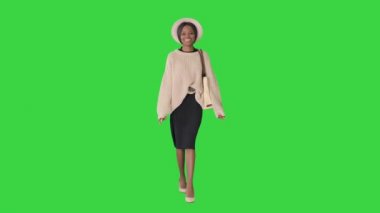 Örgü ve beyaz şapkalı gülümseyen Afrikalı Amerikalı kadın yeşil ekranda yürürken poz veriyor, Chroma Key.