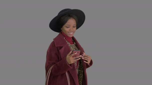 Hermosa chica de moda afroamericana en abrigo y sombrero negro mensajes de texto en su teléfono mientras camina, Alpha Channel — Vídeo de stock