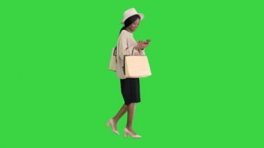 Büyük boy kazak ve şapkalı mutlu Afrikalı Amerikalı moda kızı yeşil ekranda yürürken telefonuna mesaj atıyor, Chroma Key.