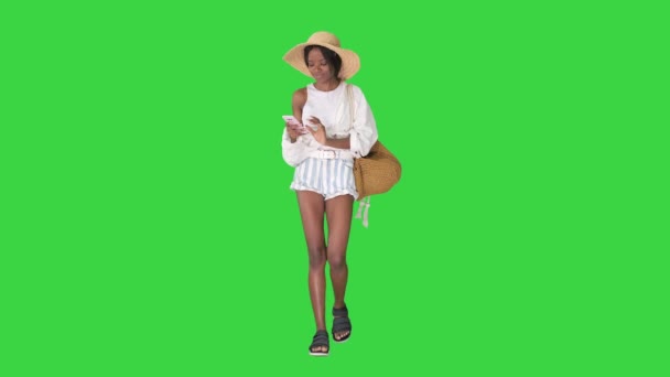 Mujer afroamericana bastante en un sombrero de paja mensajes de texto en su teléfono mientras camina en una pantalla verde, Chroma Key. — Vídeos de Stock