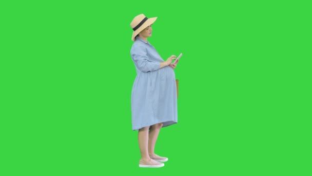 Mavi elbiseli ve şapkalı mutlu hamile kadın akıllı telefonunu yeşil ekranda kullanıyor, Chroma Key.. — Stok video