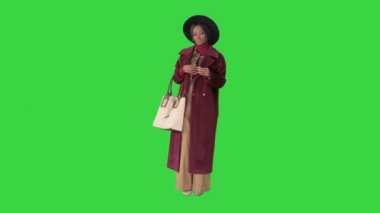 Siyah şapkalı ve ceketli Afro-Amerikan moda kızı yeşil ekranda bir çantayla poz veriyor..