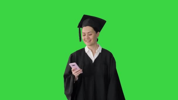 緑の画面、クロマキーの上を歩いている間、彼女の携帯電話上の段ボールテキストで笑顔の女性卒業生. — ストック動画
