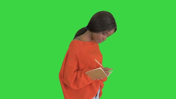 Hübsche afrikanisch-amerikanische Frau im hellen Pullover, die auf ihrem Notizblock schreibt, während sie auf einem Green Screen läuft, Chroma Key. — Stockvideo