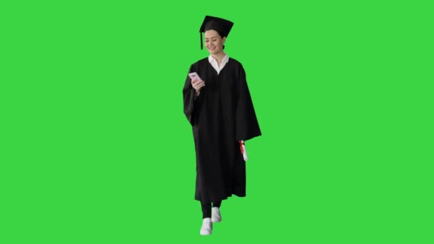 Usmívající se žena absolvent v minometu textové zprávy na svém telefonu při chůzi na zelené obrazovce, Chroma Key.
