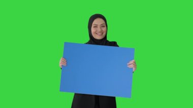 Tesettürlü gülümseyen Arap kadın elinde boş mavi posterle yeşil ekranda ona bakıyor, Chroma Key.