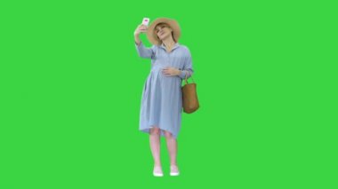 Şapkalı mutlu hamile kadın akıllı telefonunu kullanıp yeşil ekranda selfie çekiyor, Chroma Key..