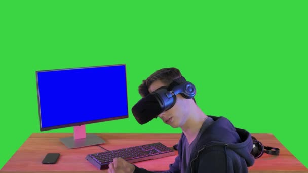Молодой игрок в наушниках виртуальной реальности сидит за столом на зеленом экране, Chroma Key. — стоковое видео