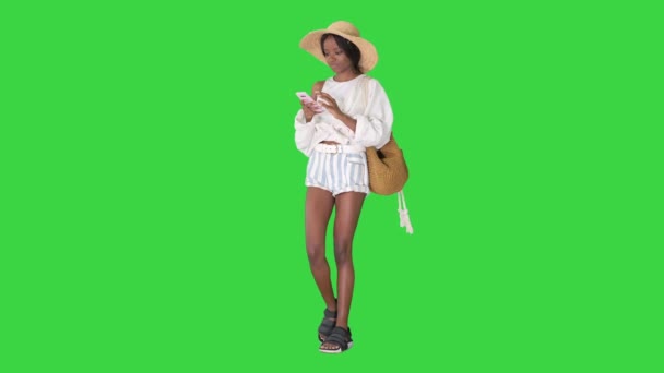 Χαμογελώντας νεαρή Αφροαμερικανή με ψάθινο καπέλο που στέλνει μηνύματα στο κινητό της σε μια πράσινη οθόνη, το Chroma Key. — Αρχείο Βίντεο