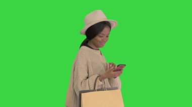 Büyük boy kazak ve şapkalı mutlu Afrikalı Amerikalı moda kızı yeşil ekranda yürürken telefonuna mesaj atıyor, Chroma Key.