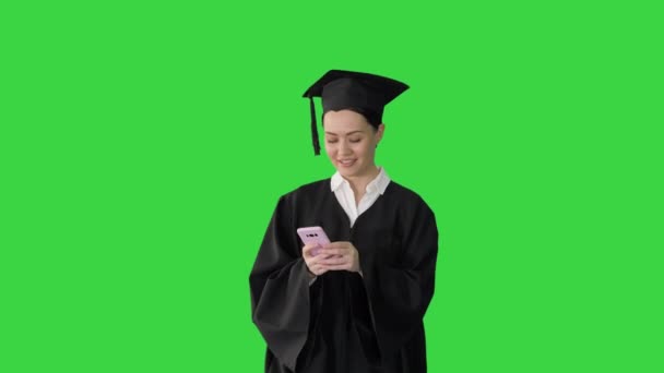 Lächelnde Absolventin in Mortarboard teilt glückliche Nachrichten auf ihrem Handy, während sie auf einem Green Screen läuft, Chroma Key. — Stockvideo
