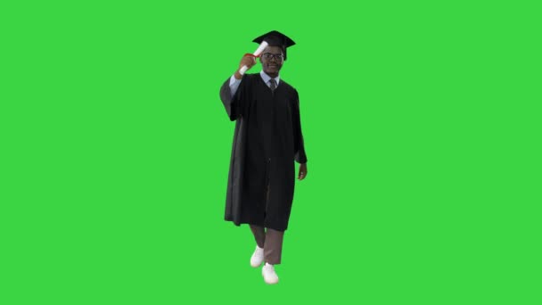Szczęśliwy afrykański student w płaszczu dyplomowym idący w kierunku kamery wymachując dyplomem na zielonym ekranie, Chroma Key. — Wideo stockowe