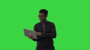 Afro-Amerikalı iş adamı dizüstü bilgisayarda yazı yazıyor ve yeşil ekranda Chroma Key 'i işaret ederken kameraya gülümsüyor..