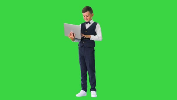 Улыбающийся мальчик в галстуке-бабочке и жилете, используя ноутбук, стоя на зеленом экране, Chroma Key. — стоковое видео