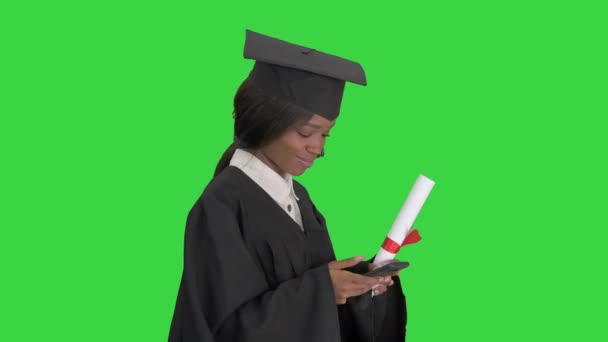 Glimlachende Afro-Amerikaanse vrouwelijke afgestudeerde met diploma sms 'en op haar telefoon tijdens het lopen op een groen scherm, Chroma Key. — Stockvideo