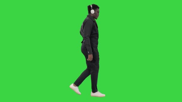 緑の画面、クロマキーを歩いている間にヘッドフォンで音楽を聴いている若いアフリカ系アメリカ人の男性. — ストック動画