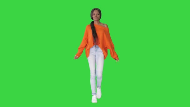 明るいセーターに身を包んだアフリカ系アメリカ人女性の笑顔とジーンズが緑の画面で彼女の髪に触れて歩く、クロマキー. — ストック動画