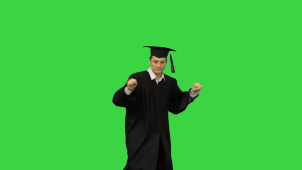 Man afgestudeerd in badjas en mortierbord doet moderne dans met armen in de lucht op zoek naar camera op een groen scherm, Chroma Key. — Stockvideo