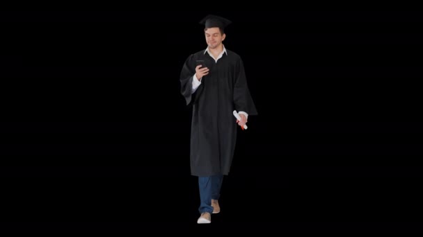 Чоловічий учень у випускній сукні перевіряє телефон під час прогулянки Альфа Канал. — стокове відео
