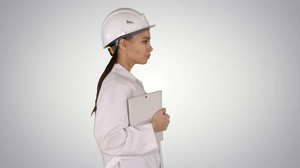 Attraktive hispanische Frau in weißem Laborkittel und weißem Schutzhelm, die mit Notizbuch oder Tablet auf Gradienten-Hintergrund geht. — Stockfoto
