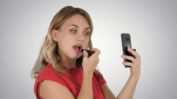 Mujer rubia aplicando lápiz labial mirando en el teléfono sobre fondo degradado. — Foto de Stock