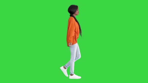 Χαμογελώντας Αφροαμερικανή γυναίκα ντυμένη με φωτεινό πουλόβερ και τζιν περπατώντας αγγίζοντας τα μαλλιά της σε μια πράσινη οθόνη, Chroma Key. — Αρχείο Βίντεο
