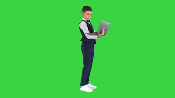 Koncentrerad pojke i fluga och väst med bärbar dator stående på en grön skärm, Chroma Key. — Stockvideo