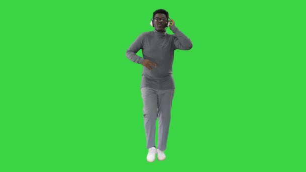 Sorrindo afro-americano homem segurando fones de ouvido com uma mão e grooving à música enquanto caminha para a câmera em uma tela verde, Chroma Key. — Vídeo de Stock