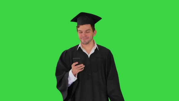 緑の画面、クロマキーを歩いている間に彼の電話をチェック卒業ガウンの男性学生. — ストック動画