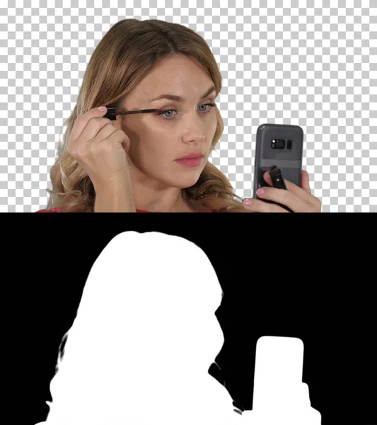 Mujer aplicando rímel negro en las pestañas mirando en su teléfono, — Foto de Stock