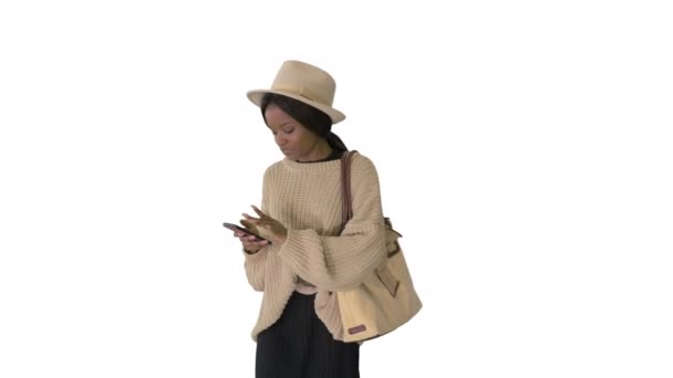 Stijlvolle Afrikaanse vrouw in gebreide kleding en hoed met behulp van haar telefoon tijdens het lopen op witte achtergrond. — Stockvideo