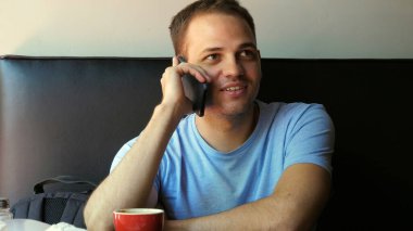 Kapalı alanda gülümseyen genç bir adamın cep telefonuyla konuşması. Kafedeki genç adam telefonda konuşuyor..